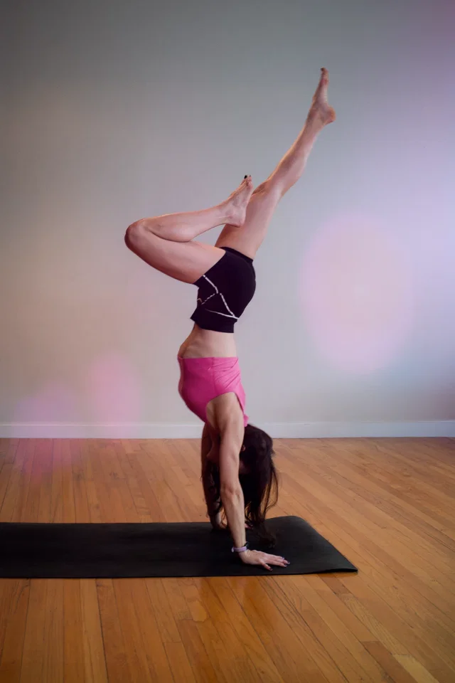 Rachael in a handstand with her legs split, on a mat on a wooden studio floor.